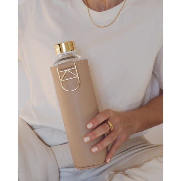 Glasflasche mit faux-leather Schutzhülle in der Farbe Ocker Beige uni. mit goldenem Tragehenkel. Flaschenform zylindrisch minimalistisch zeitlos.